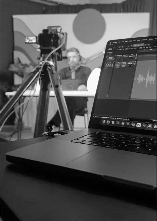 Record day: a laptop showing an audio waveform in the foreground, the host at the desk behind a camera.