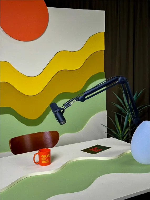 The finished set: orange sun, yellow and green waves, a microphone on a boom arm and an Okay, But... Birds mug.